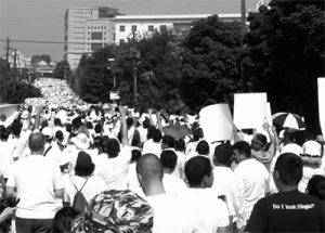 SOCI 461 / Immigration Politics and Policies B&W photo of a march for immigrant rights, with heads and raised fists down 2-3 city blocks.  In the lower right corner, a man wears a shirt that reads "Do I look illegal?"
